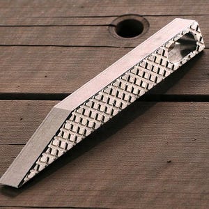 May include: A silver metal rasp tool with a textured, diamond-patterned surface. The rasp has a beveled edge and a rectangular shape, designed for shaping wood or other materials. The tool is shown on a wooden surface.
