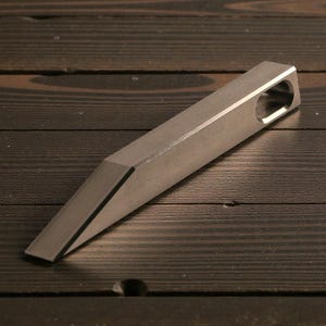 May include: A silver-colored, rectangular bottle opener with a tapered end and a hole at the opposite end. The opener is made of metal and has a matte finish. The object is angled on a dark wood surface.