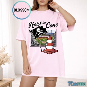 May include: Light pink t-shirt with the text "Hoist the Cone" and a graphic of a pirate flag with a skull and crossbones, a traffic cone, and a baseball field. The shirt has short sleeves and a crew neck.