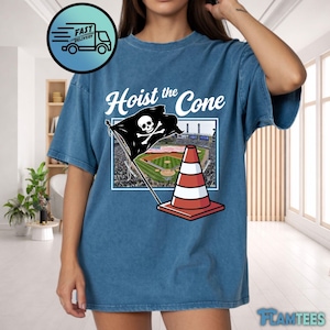 May include: Blue t-shirt with the text "Hoist the Cone" above a graphic of a pirate flag with a skull and crossbones, a baseball field, and a traffic cone. The shirt has a vintage, washed look.