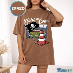 May include: Espresso-colored t-shirt with the phrase "Hoist the Cone" and a graphic of a pirate flag with a skull and crossbones, a traffic cone, and a baseball field. The shirt is a relaxed fit.