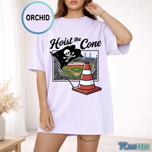 May include: A light purple t-shirt with the text "Hoist the Cone" above a graphic of a pirate flag with a skull and crossbones, a baseball field, and a traffic cone. The shirt has short sleeves.