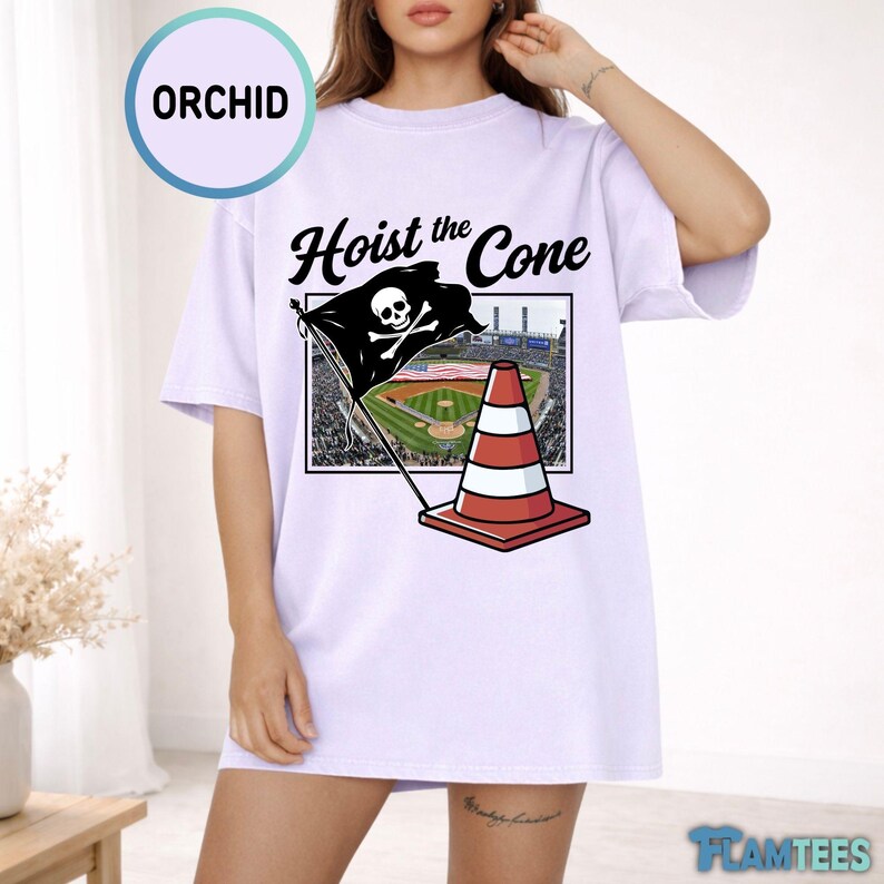 May include: A light purple t-shirt with the text "Hoist the Cone" above a graphic of a pirate flag with a skull and crossbones, a baseball field, and a traffic cone. The shirt has short sleeves.