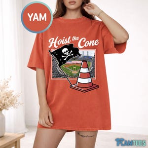 May include: Orange t-shirt with the text "Hoist the Cone" and a graphic of a pirate flag with a skull and crossbones, a traffic cone, and a baseball field. The shirt has short sleeves.