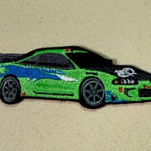 Könnte beinhalten: Gestickter Aufnäher eines grün-blauen Sportwagens mit schwarzer Motorhaube und Spoiler. Das Auto hat schwarze Räder und ein stilisiertes Design. Der Aufnäher befindet sich auf beigem Hintergrund.