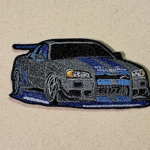 May include: Embroidered patch featuring a stylized sports car in shades of black, gray, and blue. The car has a spoiler and racing stripes. The patch is on a beige background.