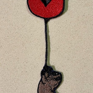 Puede incluir: Parche bordado con un círculo rojo con un corazón negro, conectado por una línea negra a una mano marrón. El parche tiene un borde negro, adecuado para ropa o accesorios.