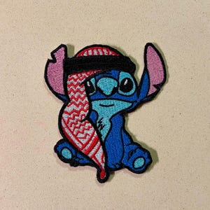 May include: Embroidered patch featuring the blue alien Stitch from Lilo & Stitch, wearing a red and white patterned scarf and a black and red head covering. The patch has a black border and is set against a neutral background.