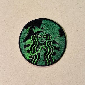 Puede incluir: Parche bordado con una figura verde con aspecto de alienígena con pelo largo y ondulado, dentro de un círculo verde con borde negro. El diseño recuerda a un logotipo de cafetería popular, con un giro único y de otro mundo.