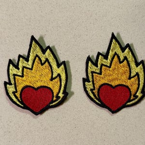 Può includere: Due toppe ricamate con un disegno a fiamma, ciascuna con un cuore rosso al centro. Le fiamme sono gialle con un contorno nero. Queste toppe sono probabilmente per abbigliamento o accessori.