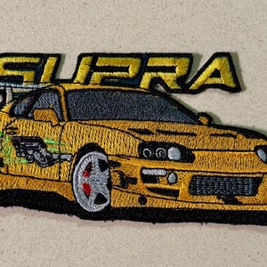 May include: Embroidered patch featuring a yellow sports car with the word "SUPRA" in yellow block letters. The car has a black spoiler, silver wheels, and green and black detailing. The patch is on a beige background.