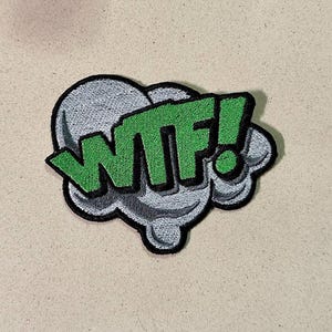 May include: Embroidered patch featuring the text "WTF!" in bright green, outlined in black, set against a gray cloud-like shape. The patch has a black border and is likely designed to be ironed or sewn onto fabric.