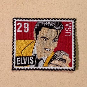May include: Embroidered patch depicting Elvis Presley as a postage stamp. The patch has a red background with the text "USA" and "29". Elvis is shown in a yellow jacket, holding a microphone, with the name "ELVIS" below.