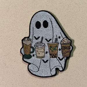 May include: Embroidered patch of a ghost holding four coffee drinks. The ghost is light grey with black eyes and small bats. The coffee cups are brown and beige with whipped cream and straws.