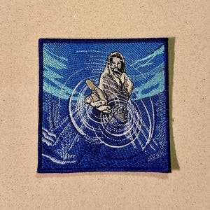 May include: Embroidered blue square patch featuring a figure reaching out, encircled by concentric white lines. The figure is rendered in brown and white tones, set against a blue and teal backdrop.