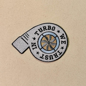 May include: Embroidered patch with a turbocharger design. The gray patch has black trim and the text "IN TURBO WE TRUST" encircling a fan design with blue, gold, and brown details. The patch is set against a tan background.