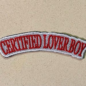 May include: Embroidered patch with the text "CERTIFIED LOVER BOY" in red, outlined in white, and bordered with black stitching. The patch has a curved shape and is set against a neutral, textured background.