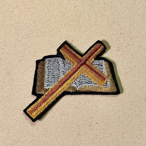 May include: Embroidered patch featuring a gold and red cross laid over an open book. The book has silver-colored pages and a gold border. The patch has a black border and is likely for clothing or accessories.