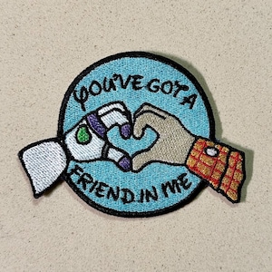 May include: Embroidered patch featuring two hands forming a heart shape. One hand is a white robotic arm, the other is a tan human hand. The patch is round with the text "YOU'VE GOT A FRIEND IN ME".