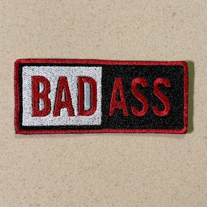 Può includere: Toppa ricamata con le parole "BAD ASS" in filo rosso. Il lato sinistro della toppa è bianco e il lato destro è nero. La toppa ha un bordo rosso.