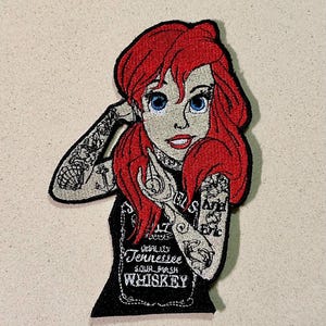 Può includere: Toppa ricamata raffigurante una sirena dei cartoni animati con capelli rossi e occhi blu, ornata di tatuaggi. Indossa un top nero con la scritta "Tennessee Sour Mash Whiskey". La toppa è su sfondo beige.