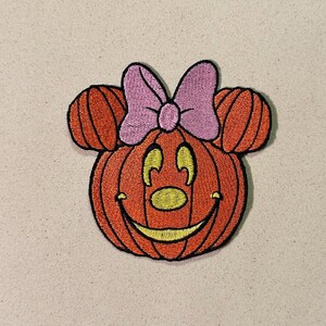 以下が含まれることがあります： マウスの耳とピンクのリボンが付いた、笑顔のカボチャの刺繍パッチ。カボチャはオレンジ色で、黄色い鼻と口があり、黒で縁取られています。楽しいハロウィーンをテーマにしたパッチです。