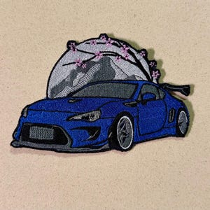 Puede incluir: Parche bordado con un coche deportivo azul con un alerón negro. El coche está situado sobre un fondo de montaña y ramas de flores de cerezo. El parche está sobre una tela beige.
