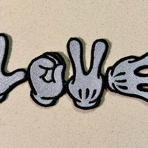 Puede incluir: Parche bordado que deletrea "LOVE" con manos de estilo caricatura. Las letras están formadas por las manos en gris con contornos negros. El fondo es de color beige claro.