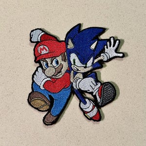Può includere: Toppa ricamata con Mario e Sonic the Hedgehog. Mario indossa un cappello e una tuta rossa, mentre Sonic è blu. Entrambi i personaggi sono in pose dinamiche, con cuciture dettagliate e un bordo nero.