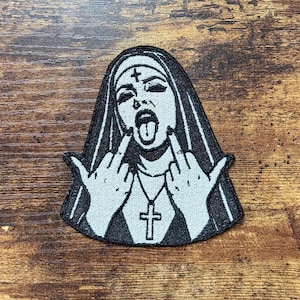 May include: A black and white embroidered patch featuring a nun wearing a habit with a cross on the forehead, a cross necklace, and dark makeup. The figure has an open mouth with the tongue out and is gesturing with both middle fingers.