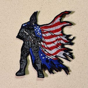 May include: Embroidered patch featuring a soldier silhouette with a tattered American flag design. The soldier is depicted in black and gray, holding a rifle. The flag displays red and white stripes and blue stars.