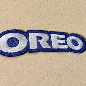Op de afbeelding: Geborduurde patch met het woord "OREO" in wit, omlijnd door een donkerblauwe rand. De patch ligt op een beige oppervlak. De letters zijn in een vet, afgerond lettertype, wat zorgt voor een eenvoudig en herkenbaar ontwerp.
