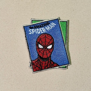May include: Embroidered patch featuring the Amazing Spider-Man comic book cover. The patch has a blue background with the title "The Amazing Spider-Man" and a red and black Spider-Man face. The patch is layered on top of a green and red patch.