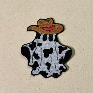 May include: Embroidered patch featuring a ghost wearing a brown cowboy hat with a red band. The ghost has a light gray body with black spots, and a black outline. This patch is suitable for jackets, bags, or other fabric items.