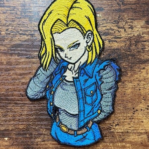 May include: An embroidered patch featuring a character with bright yellow hair, wearing a blue denim vest over a gray shirt, with gray long sleeves, a brown belt with a gold buckle, and gold hoop earrings, rendered in a detailed anime style.