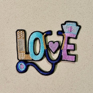 May include: Embroidered patch spelling "LOVE" with medical theme. The "L" is a bandage design, the "O" is turquoise, the "V" is a stethoscope, and the "E" is a nurse's cap. The heart is purple.