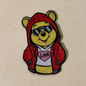 May include: Embroidered patch of a yellow bear in a red hoodie and sunglasses. The bear wears a white shirt with a pink heart and the word "Love". The patch has a black border.