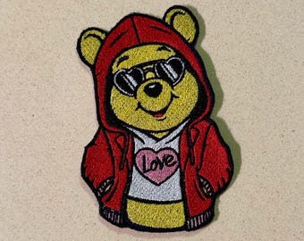 Puede incluir: Parche bordado con un oso amarillo con una sudadera roja y gafas de sol. El oso lleva una camiseta blanca con un corazón rosa que dice "Love". El parche tiene un borde negro.