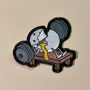 May include: Embroidered patch featuring a cracked egg character lifting weights on a bench. The egg has crossed-out eyes and yolk dripping down. The patch has a black border and is on a tan background.