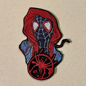 Puede incluir: Parche bordado con un diseño de Spider-Man. El parche muestra una figura roja con capucha con una máscara de telaraña en blanco y negro. Un emblema de araña roja está en la parte inferior. El parche está sobre un fondo beige.