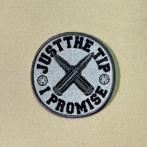 May include: Circular patch with the text "JUST THE TIP I PROMISE" in black lettering. Two crossed bullets are in the center. The patch has a gray background and a black border.