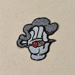 May include: Embroidered patch featuring a cartoon hand holding a lit cigar, with smoke rising above. The hand is white with black outlines, the cigar is brown, and the smoke is gray. A red ember glows at the cigar's tip.