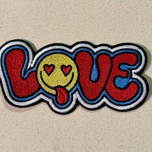Puede incluir: Parche bordado con la palabra "LOVE" en rojo, azul y blanco. La "O" es una carita sonriente amarilla con ojos de corazón y una lengua roja. El parche tiene un borde negro sobre un fondo beige.