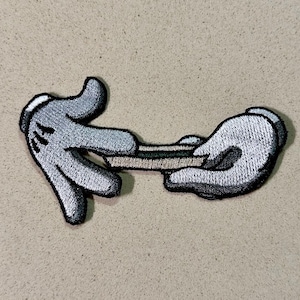 May include: Embroidered patch featuring two cartoon-style white gloves with black outlines, holding a rolled object. The gloves are gray with black details. The patch is on a tan background.