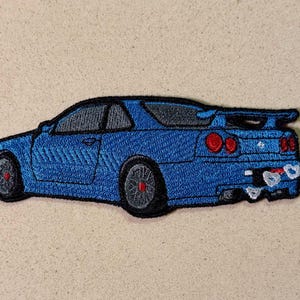 May include: Embroidered patch featuring a blue sports car with a black roof and spoiler. The car has red rear lights and grey wheels. The patch is on a beige background.