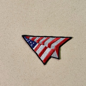 May include: Embroidered patch in the shape of a stylized American flag. The patch features red and silver stripes, a blue field with white stars, and a black border. The patch is likely for clothing or accessories.