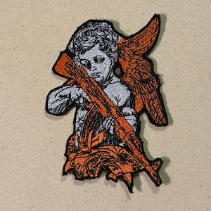 May include: Embroidered patch depicting a cherub with orange wings and an orange rifle. The patch is mainly white, with black outlining and orange details. The design is set against a tan backdrop.