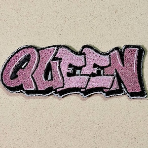 Puede incluir: Parche bordado con la palabra "QUEEN" en estilo grafiti. El texto es en tonos rosas con un contorno negro. El parche está sobre un fondo neutro. Adecuado para ropa o accesorios.