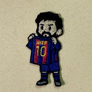 Op de afbeelding: Geborduurde patch met een cartoonfiguur die een blauw en rood voetbalshirt omhoog houdt. Het shirt toont het nummer 10 en de naam "MESSI" in gouden letters. De figuur heeft zwart haar en een baard.