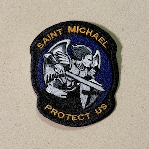 May include: Embroidered patch featuring Saint Michael, depicted with a sword and shield. The patch has a black border with the words "SAINT MICHAEL" and "PROTECT US" in gold lettering. The background is blue.
